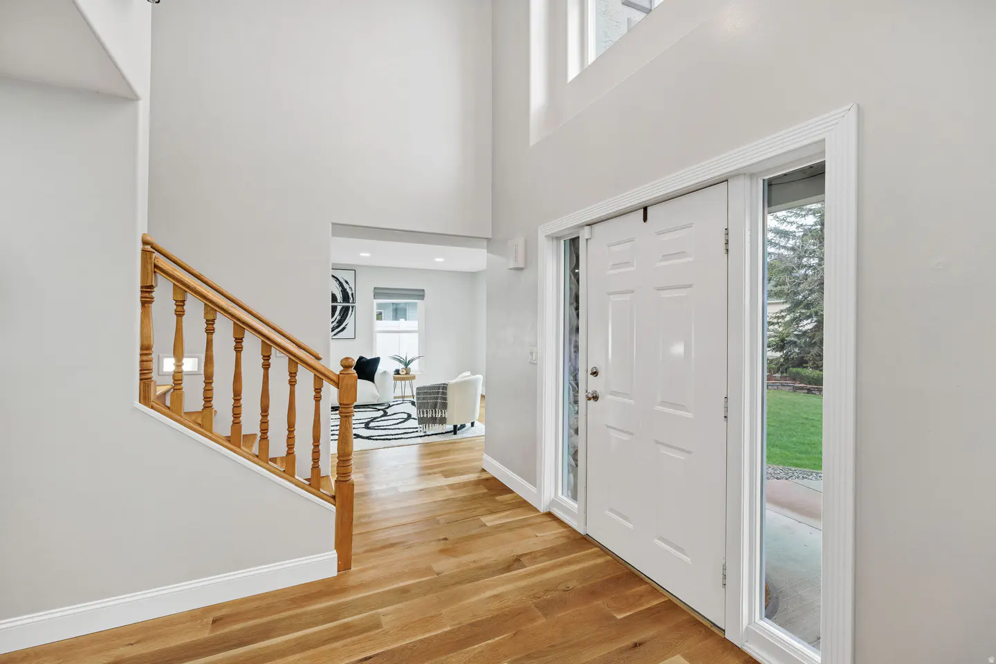 Bright foyer with hardwood floors, white walls, and a staircase with a wooden banister. A white front door with sidelights is on the right.
