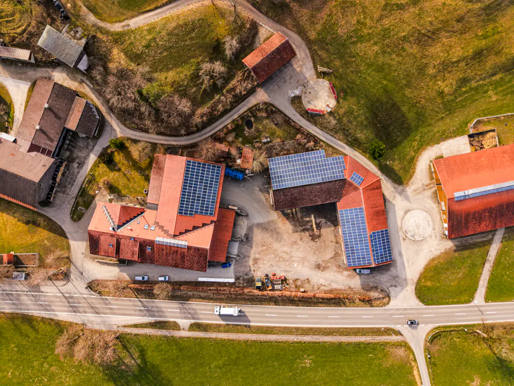Aerial view of a farm with red-roofed buildings, solar panels, green fields, and a road with a white truck.