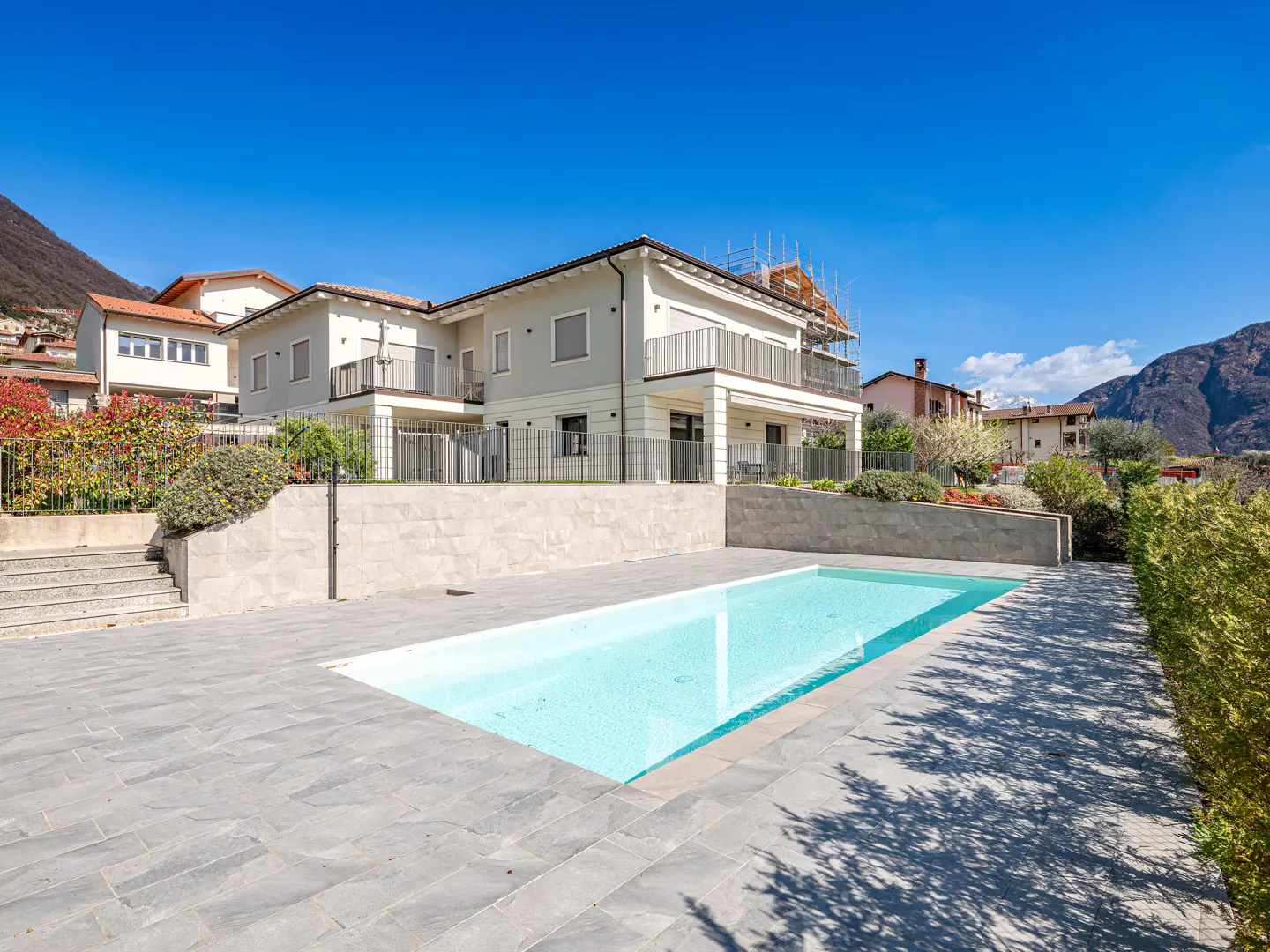 A modern two-story house with a pool, gray stone patio, and mountain backdrop under a clear blue sky.