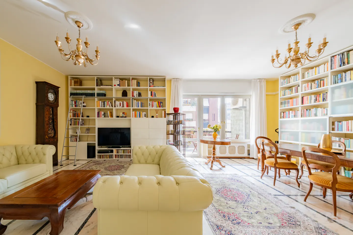 Bright living room with yellow walls, white bookshelves filled with books, and a cream-colored leather sofa. A wooden table and chairs are on a patterned rug.