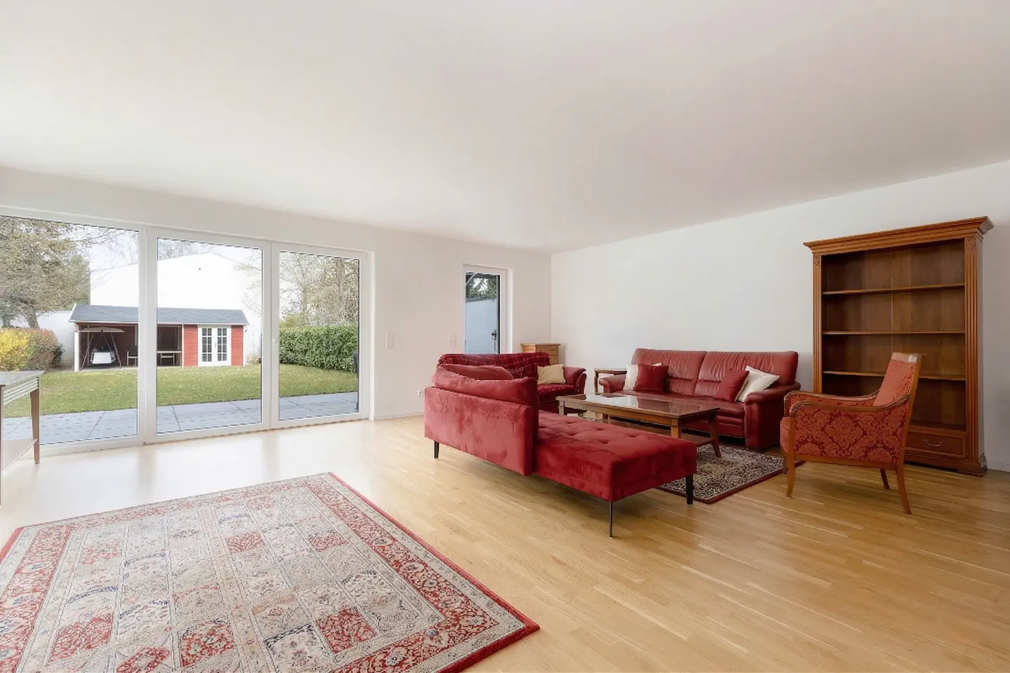 Bright living room with red sofas, wood floors, and a large window overlooking a green yard with a red shed.