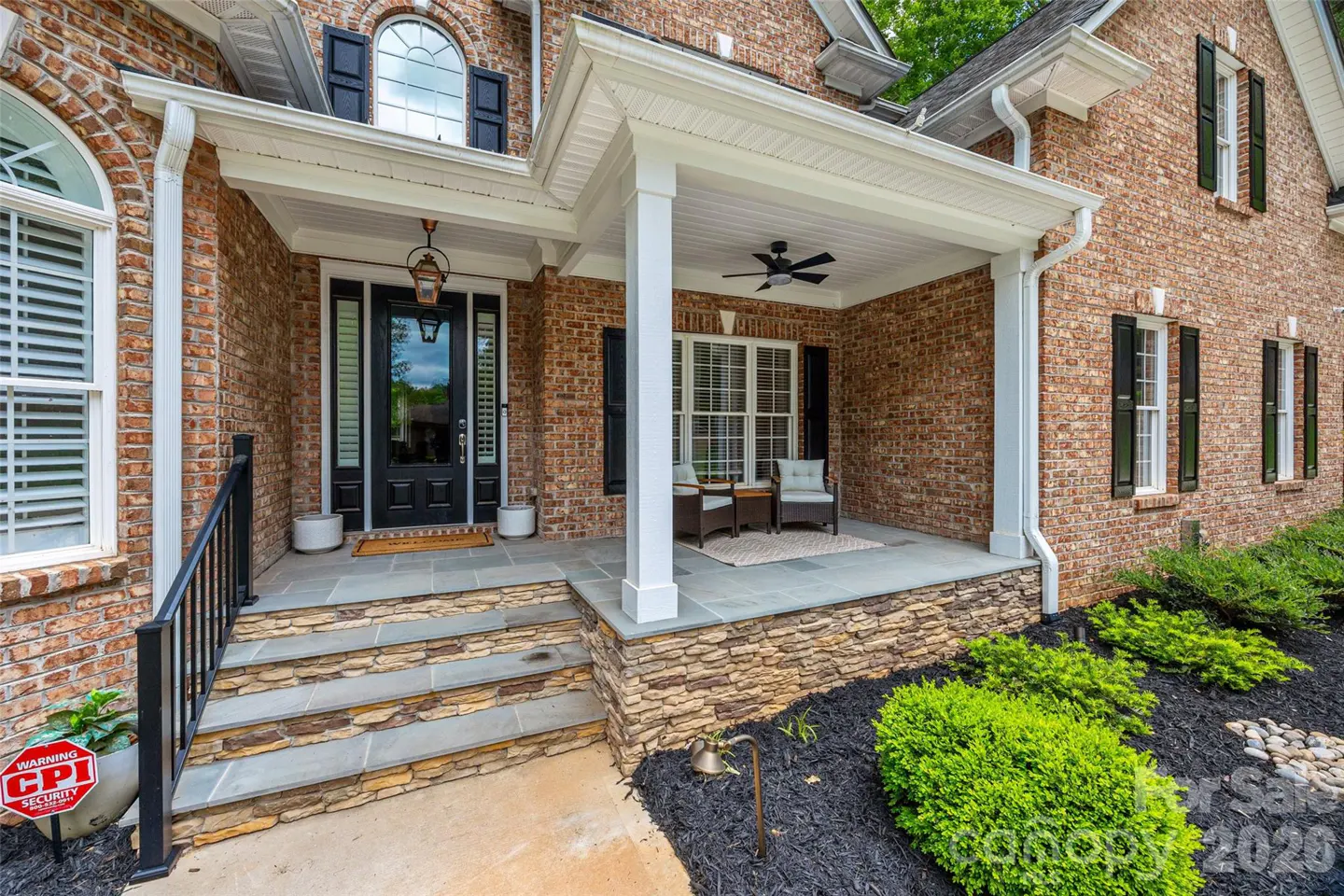 Brick home exterior with a covered front porch, stone steps, black front door, and two chairs on a landscaped lawn.