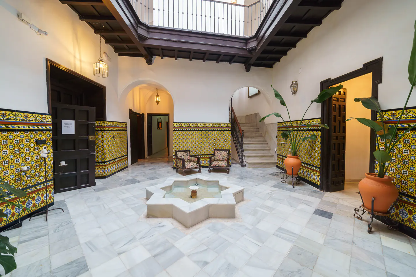 Interior courtyard with marble floors, star-shaped fountain, tiled walls, dark wood beams, and potted plants.