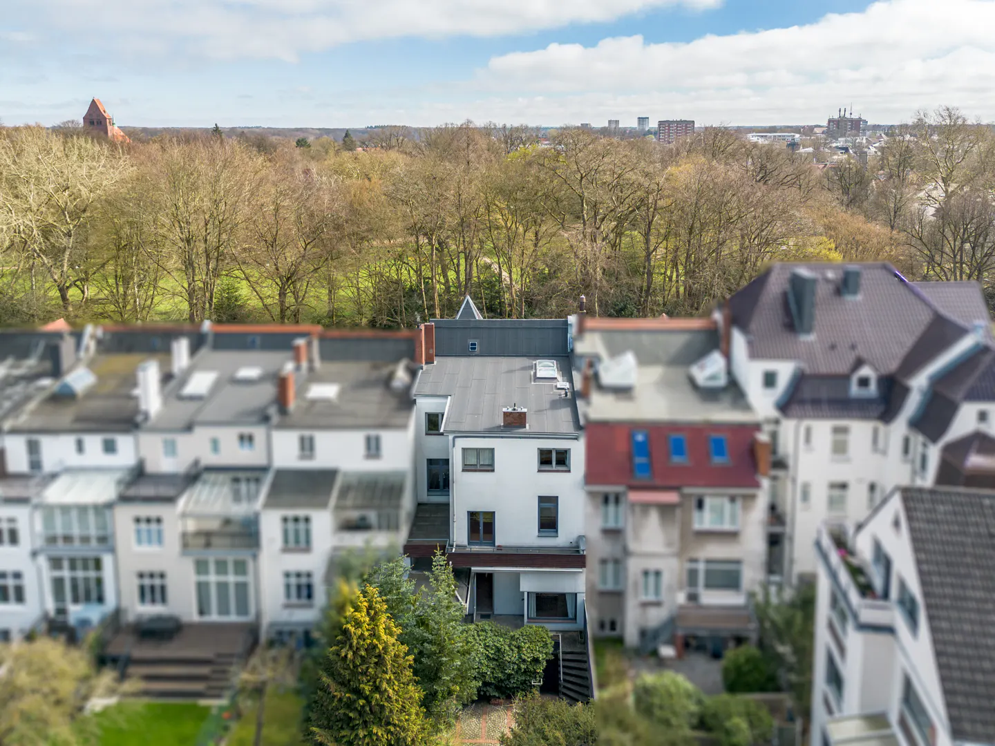 Aerial view of a white, multi-story house with a black roof, surrounded by greenery and other houses. A park and city skyline are in the background.