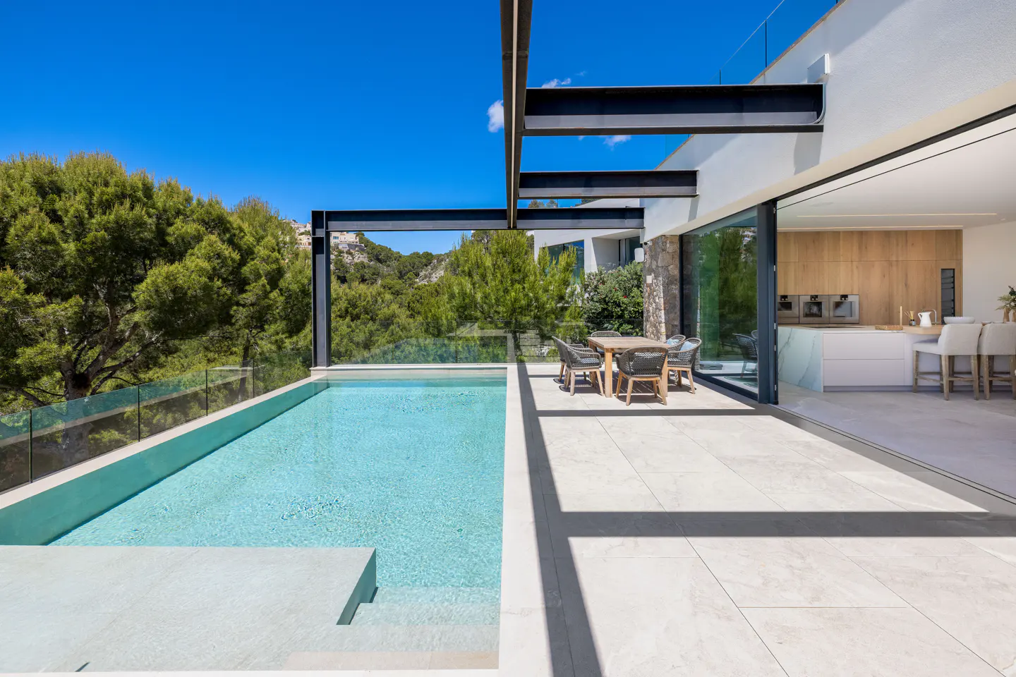Outdoor patio with pool, dining table, and open sliding doors to a modern kitchen. Blue sky and green trees in background.