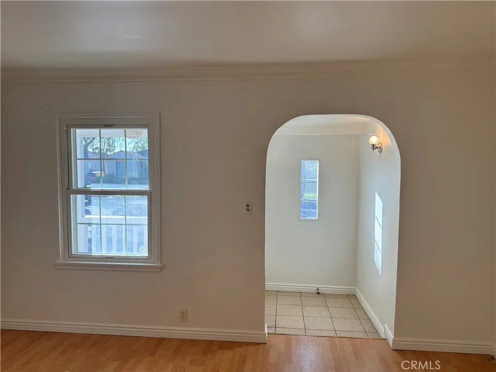 A bright room with hardwood floors, white walls, a window, and an arched doorway leading to a tiled area.
