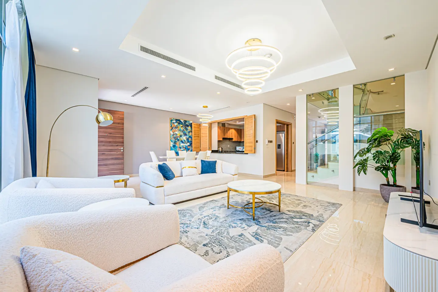 Bright, modern living room with white sofas, blue accents, and a patterned rug. A gold floor lamp and a round coffee table add elegance. Kitchen visible in the background.