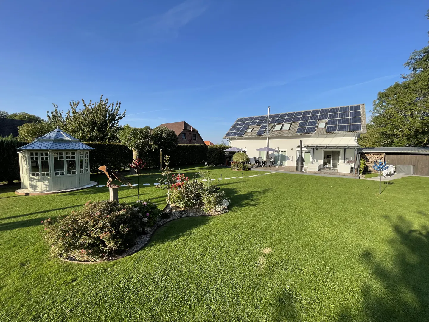 A wide shot of a green lawn, a gazebo, and a white house with solar panels under a blue sky.