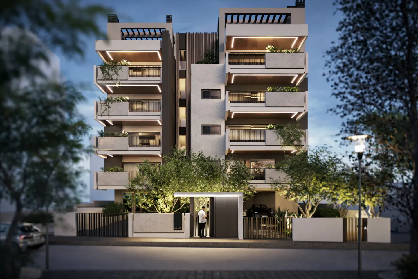 Modern apartment building with balconies and plants, lit by warm lights at dusk. A person enters the gate, with trees and a street lamp nearby.