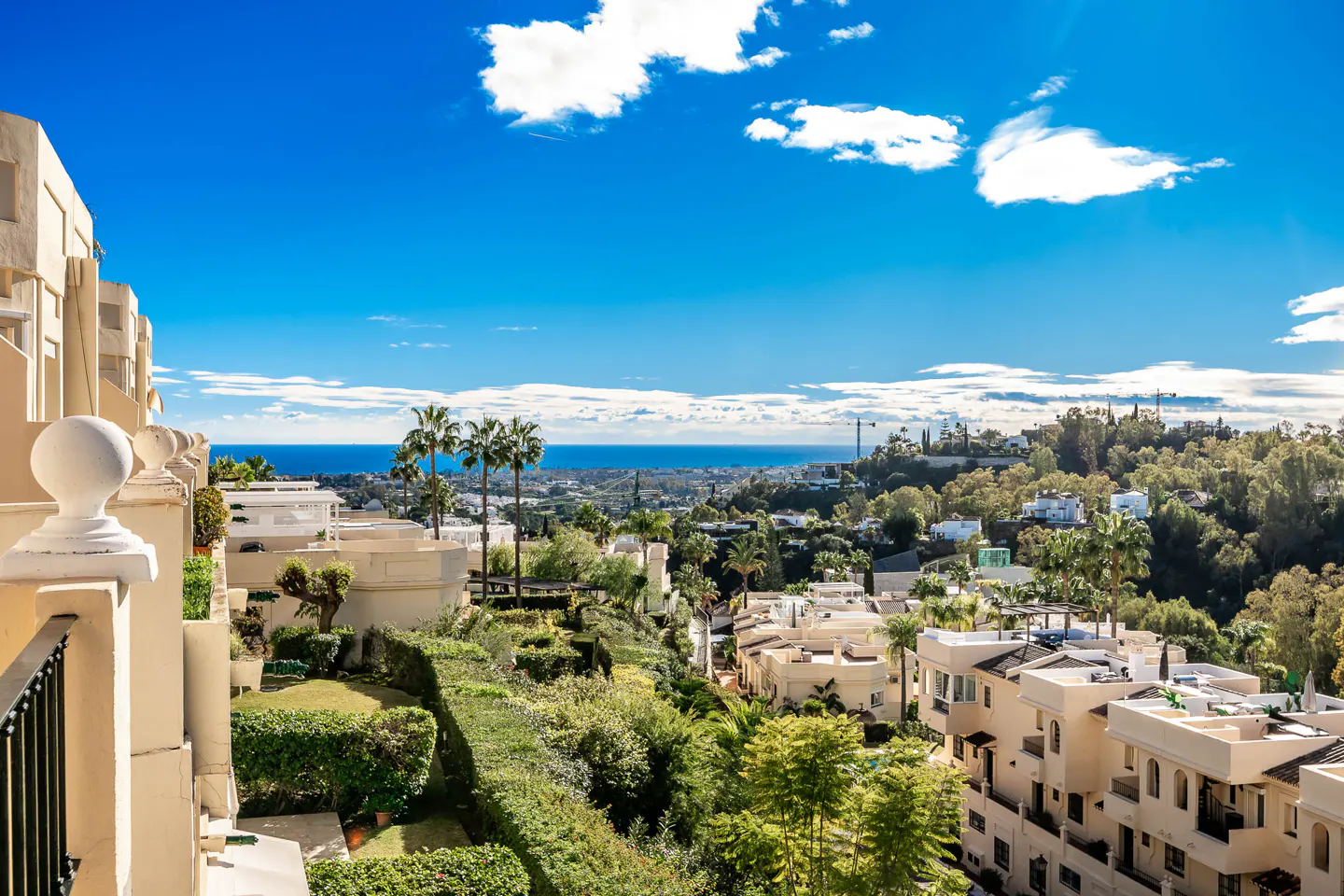 View from a balcony overlooking a lush green landscape with white buildings, palm trees, and the ocean under a blue sky.