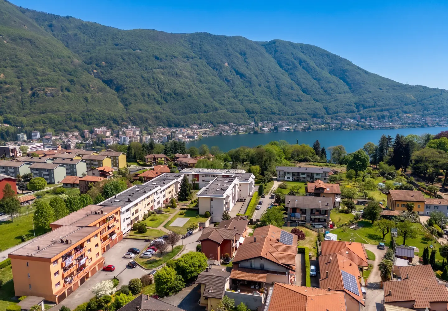Aerial view of a town with orange-roofed houses and apartments near a lake and green mountains under a clear blue sky.