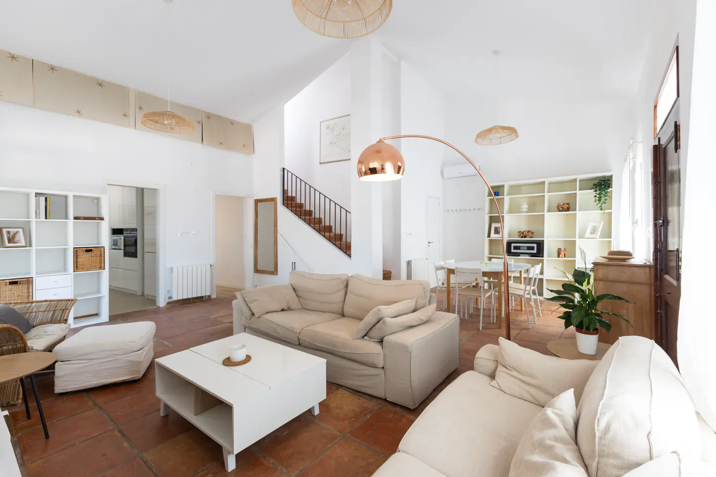 Bright living room with white walls, terracotta tile floor, and beige sofas. A white coffee table sits in front of the sofa. A staircase is visible in the background.