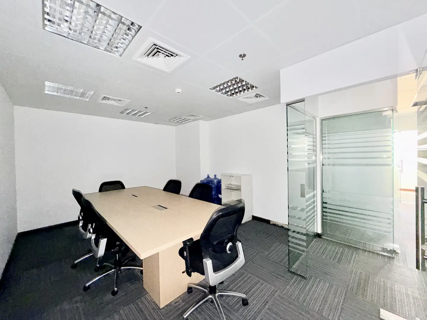 Modern office meeting room with a light wood table, black ergonomic chairs, gray carpet, and glass-walled partitions.
