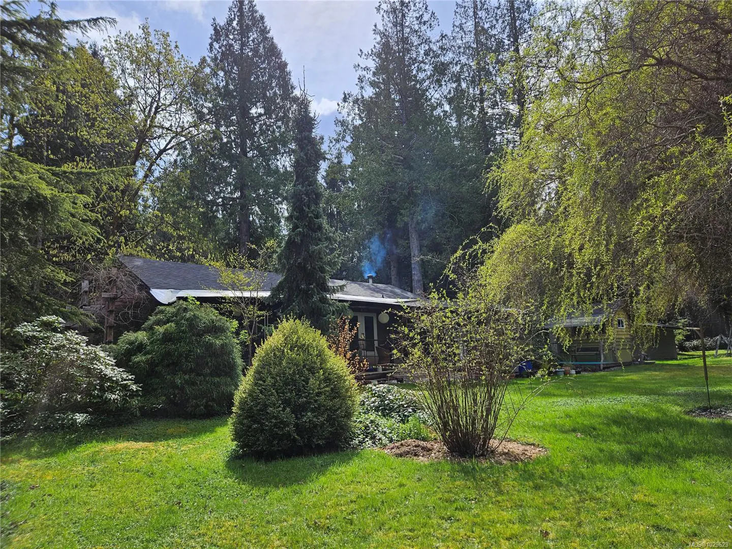 A house with a dark roof is surrounded by green trees and a lawn. Smoke rises from the chimney.