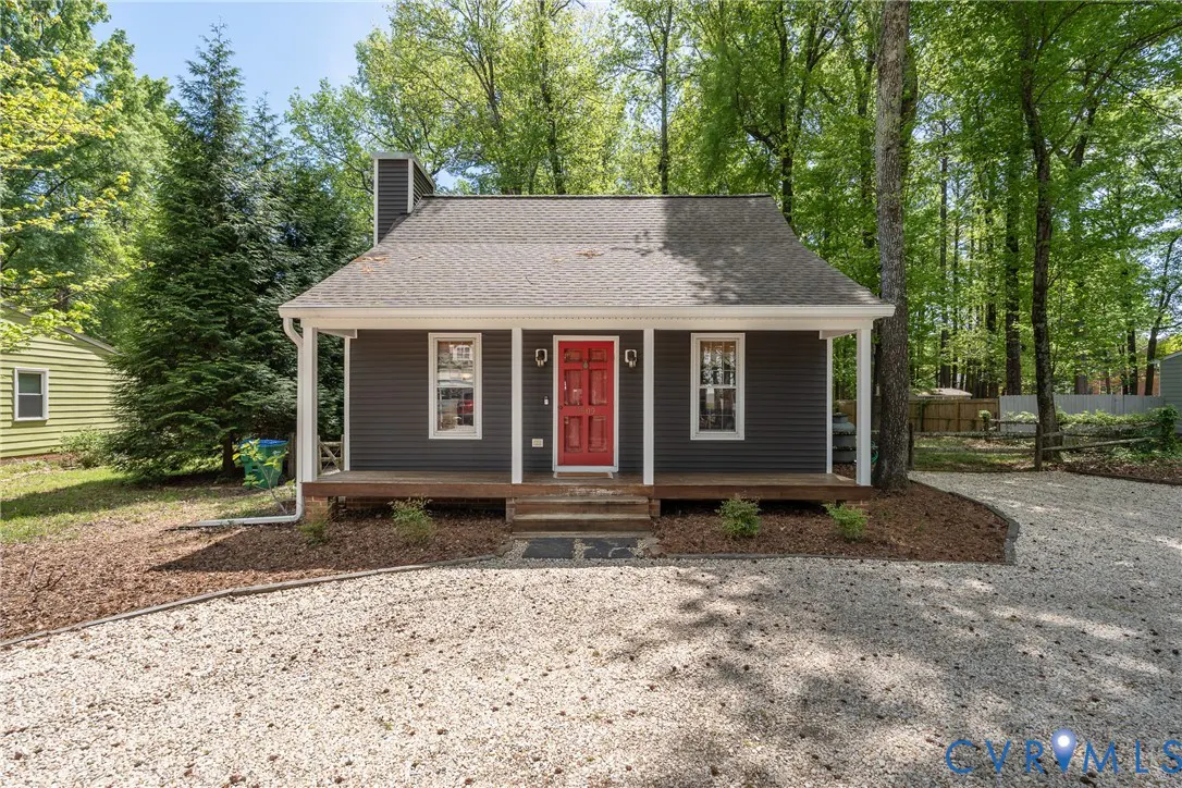Front view of a gray house with a red door, white trim, and a gravel driveway. Trees surround the house.