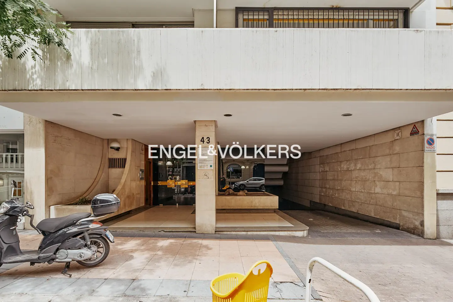 Entrance of a modern building with a stone facade, a covered driveway, and a parked scooter in the foreground.
