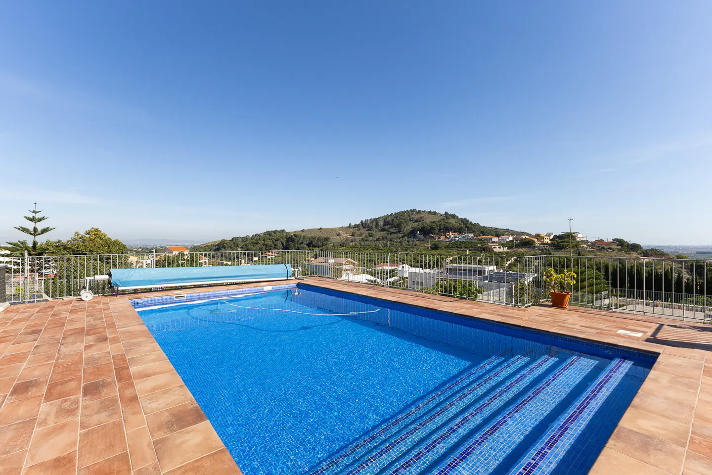 Outdoor pool with blue tiles and steps, surrounded by terracotta tiles and a metal fence, overlooking a green hillside under a clear blue sky.