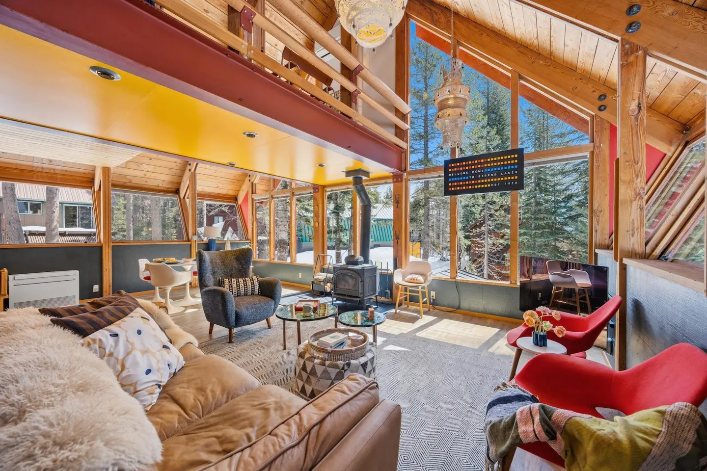 A-frame cabin interior with wood beams, large windows, and a mix of modern and rustic furniture. A wood-burning stove sits near the windows.