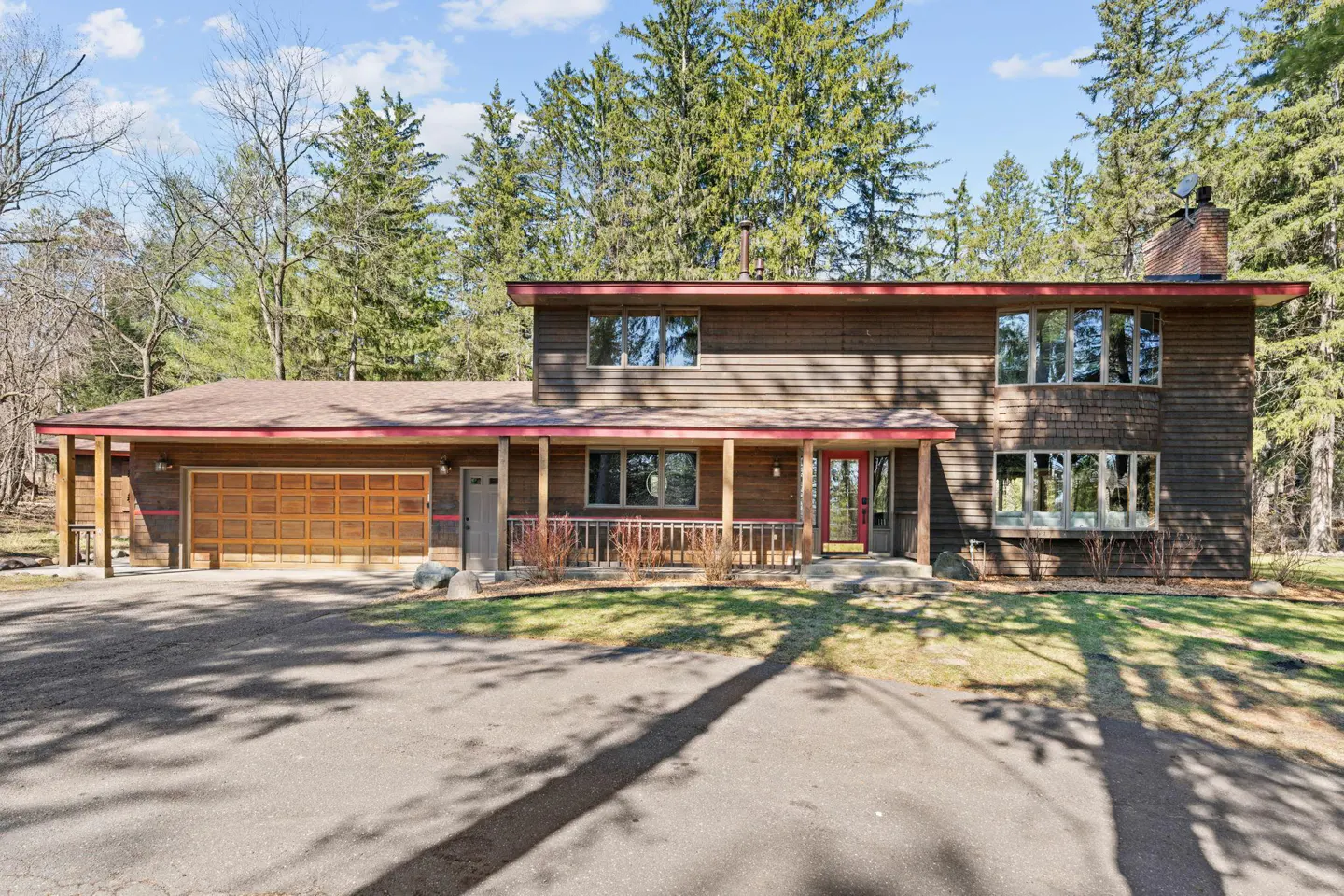 Two-story brown house with a red trim, a wooden garage door, and a chimney, surrounded by trees.
