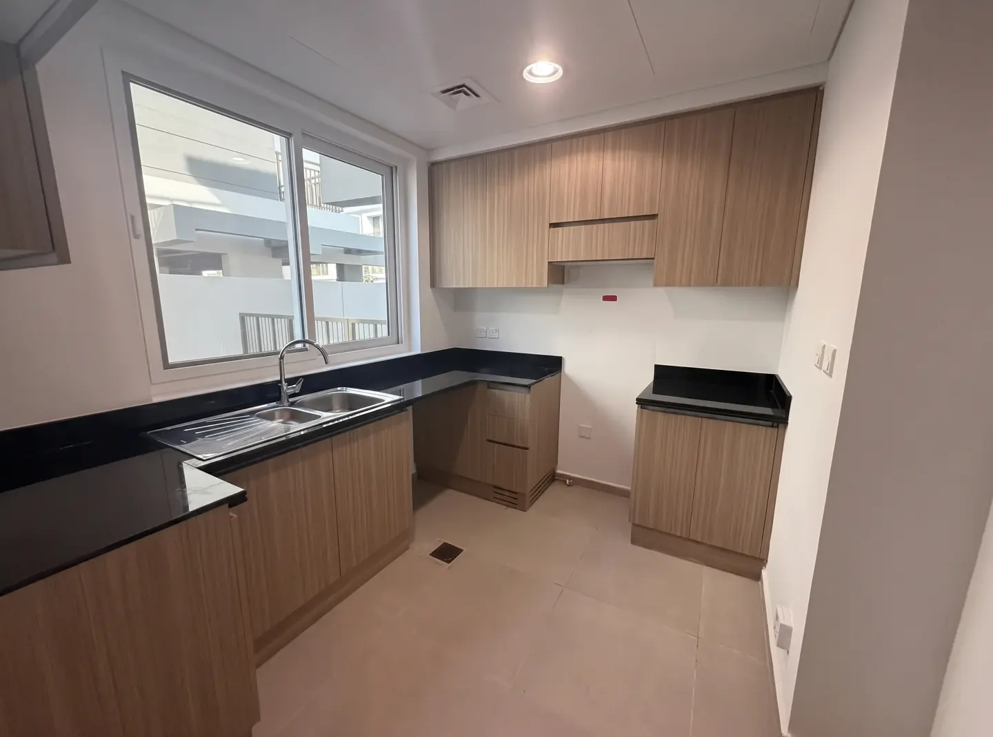 A bright, empty kitchen with light wood cabinets, black countertops, and a stainless steel sink under a window.