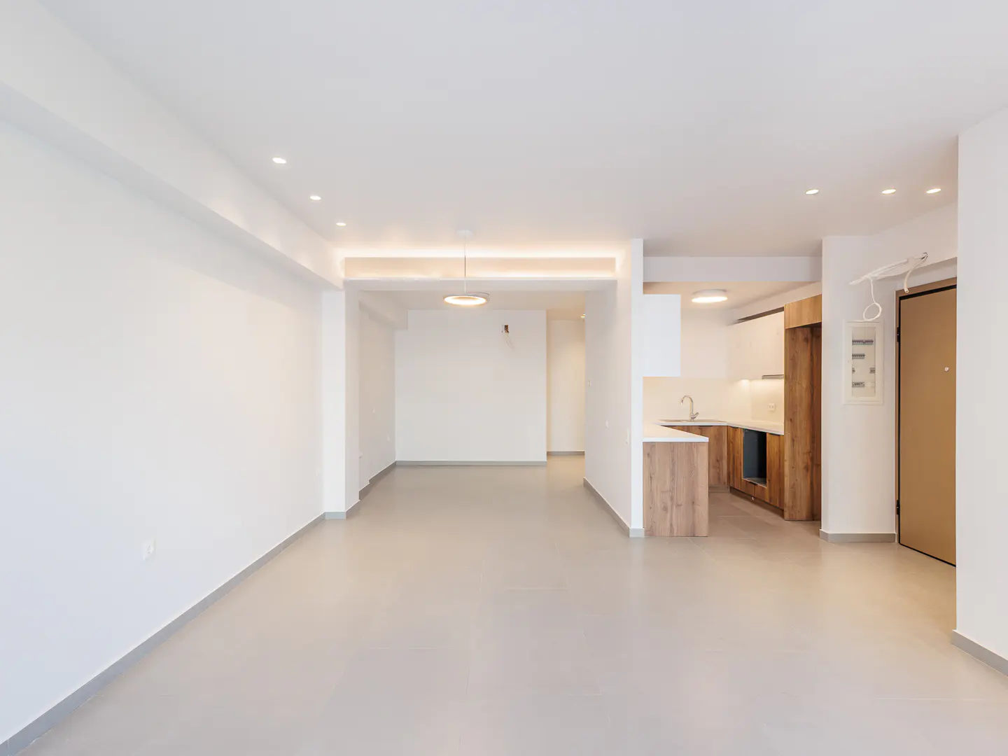 Modern open-plan living area with white walls, light grey floors, and a small kitchen with wood-toned cabinets.