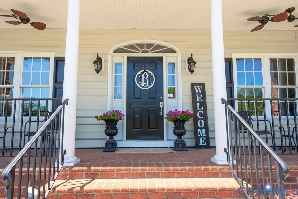 Front porch of a beige house with a black door, flanked by flower pots and a welcome sign.
