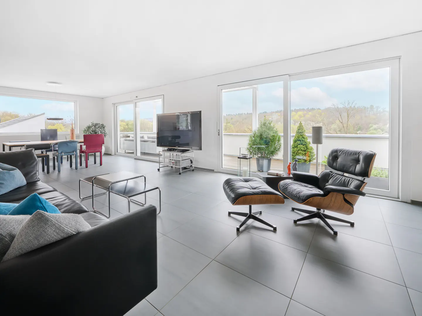 Bright, modern living room with a black leather sofa, lounge chair, TV, dining table, and large windows with a view.