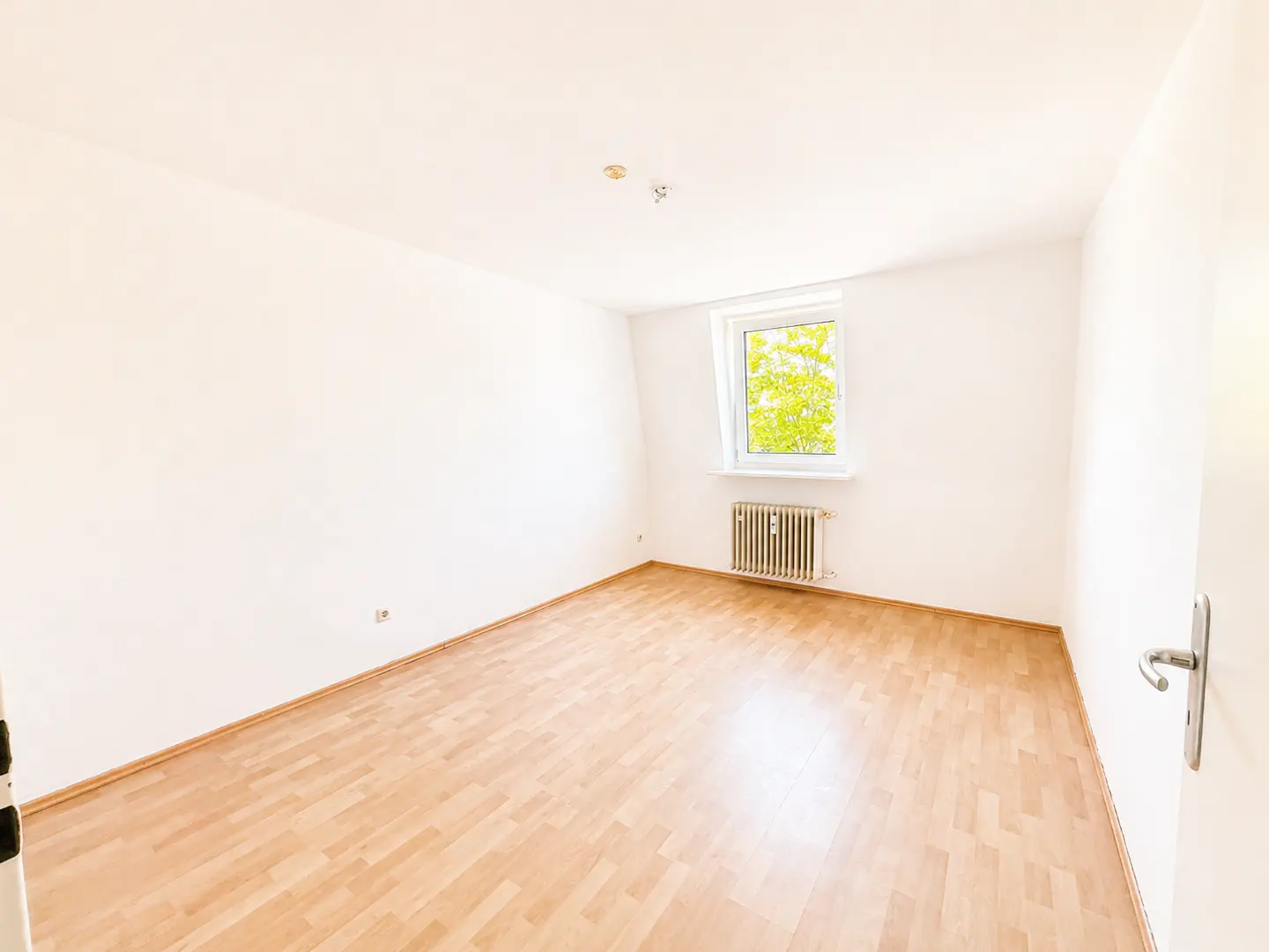 Bright, empty room with light wood floors and white walls. A window shows green trees. A radiator sits below the window.