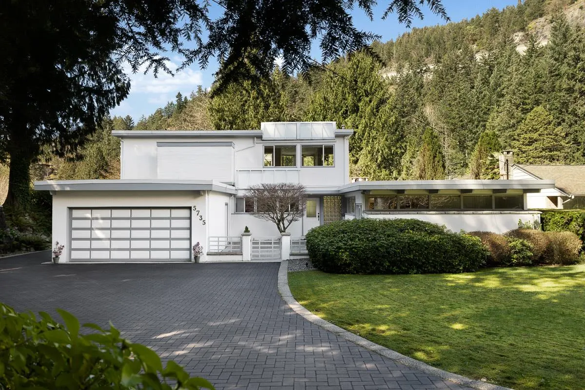 A modern white house with a gray roof and garage door sits on a green lawn with trees in the background.