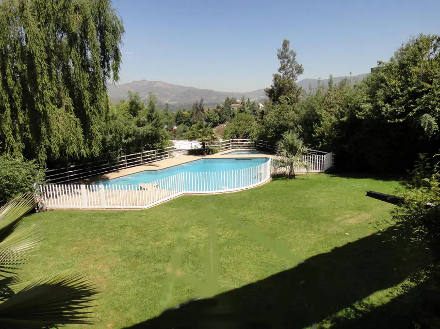 A backyard with a blue swimming pool surrounded by a white fence and green grass. Trees and mountains are in the background.