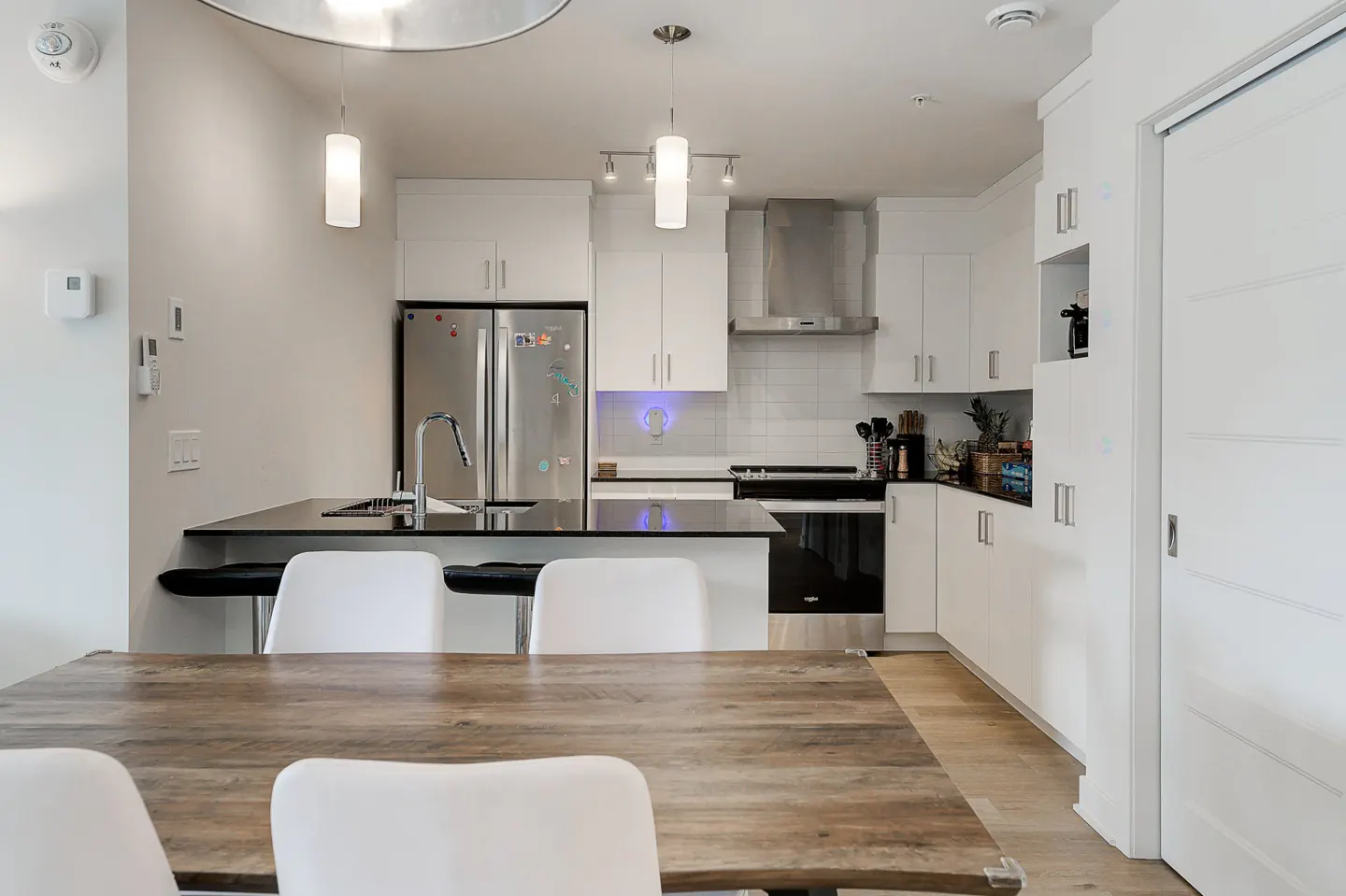 Bright kitchen with white cabinets, stainless steel appliances, and a black countertop island. A wooden dining table with white chairs is in the foreground.