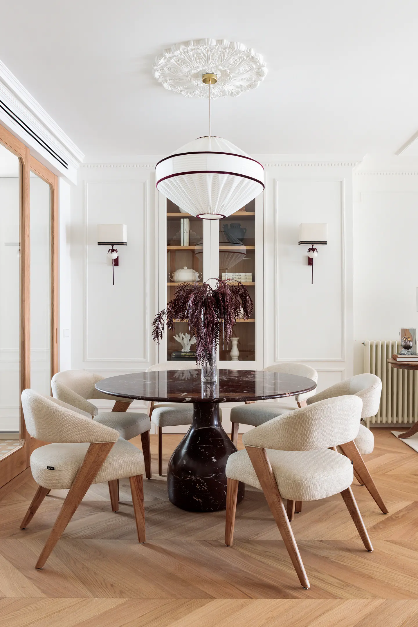 Bright dining room with a round, dark marble table, six cream chairs with wood frames, and a large white pendant light.