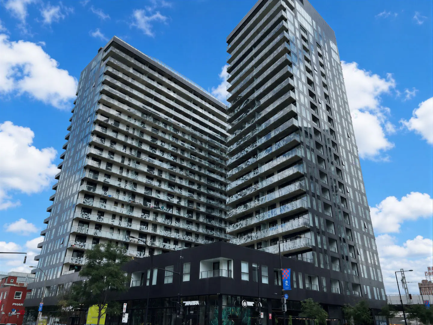 Two modern high-rise apartment buildings with balconies, under a blue sky with white clouds. Street-level retail space below.
