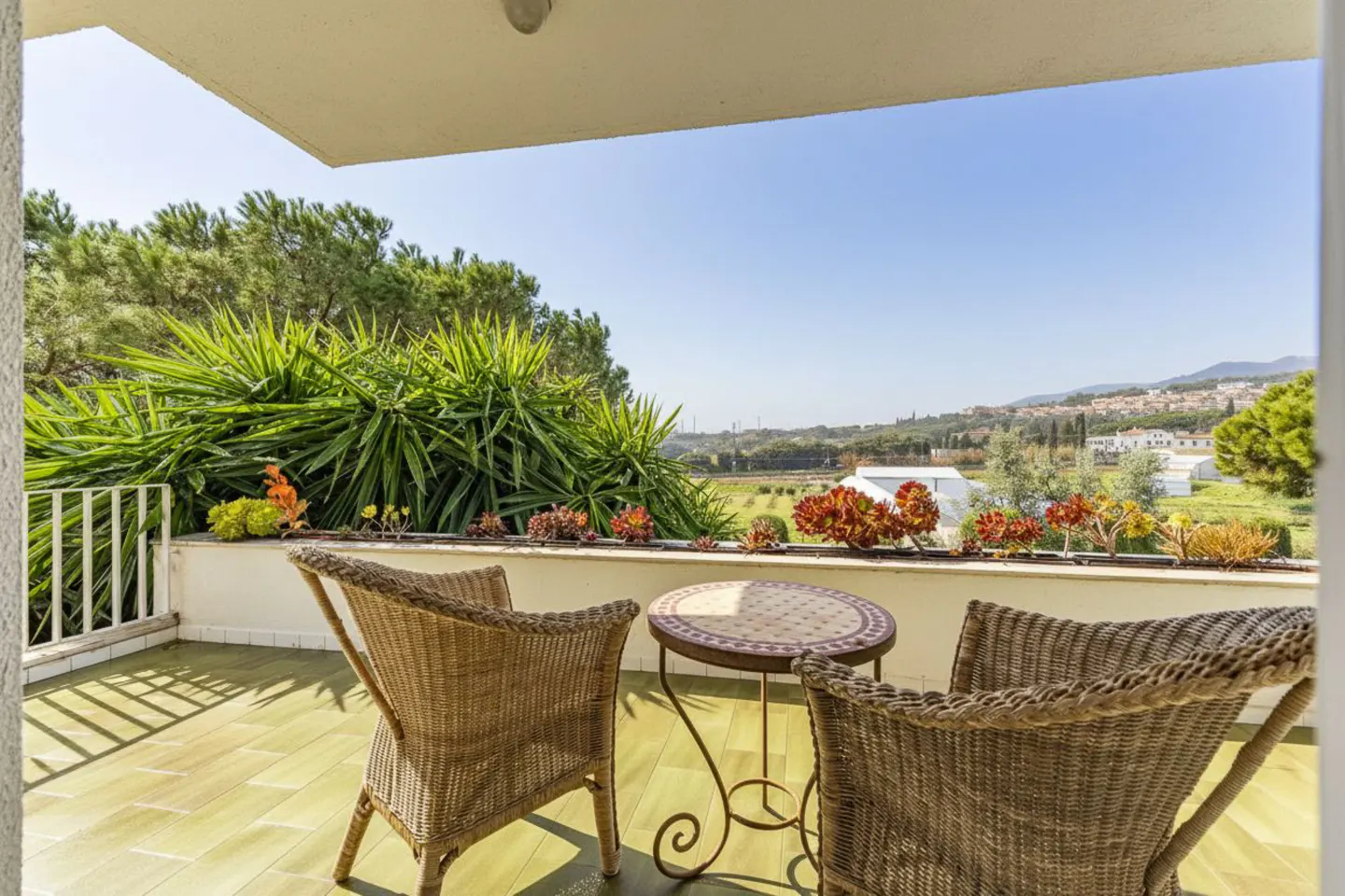 Balcony with two wicker chairs and a mosaic table overlooking a green landscape under a blue sky.