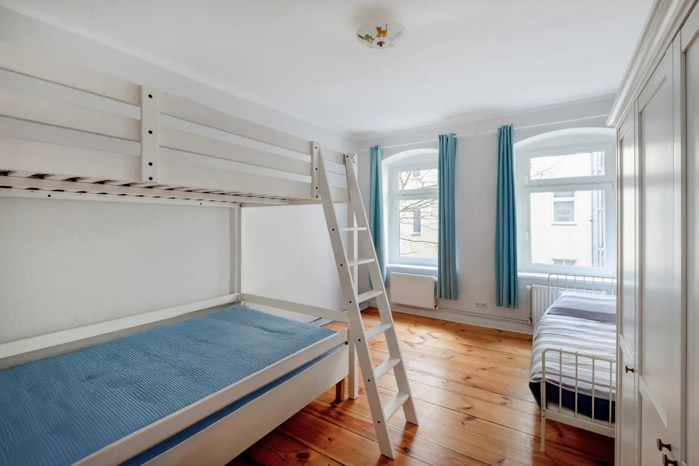 Bright bedroom with white bunk beds, a single bed, and hardwood floors. Blue curtains frame the windows.