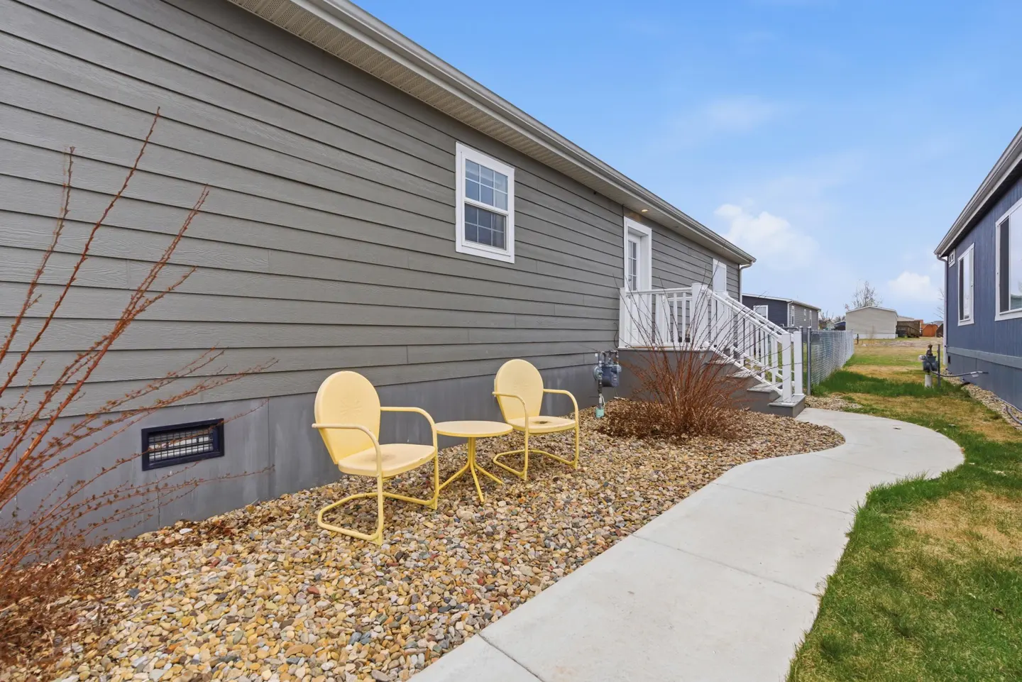 Exterior view of a gray house with yellow chairs and table on a gravel bed, next to a curved sidewalk.