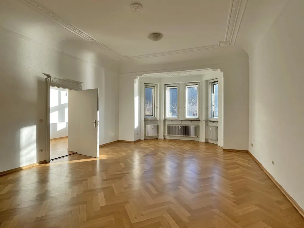 Empty room with herringbone wood floor, white walls, and bay window with radiators. Open door to another room.