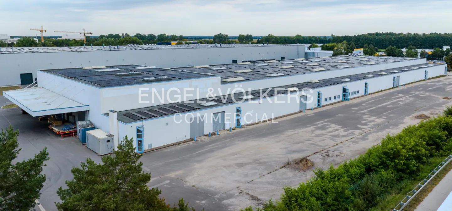 Aerial view of a long, white commercial building with solar panels on the roof, surrounded by trees and a large parking lot.