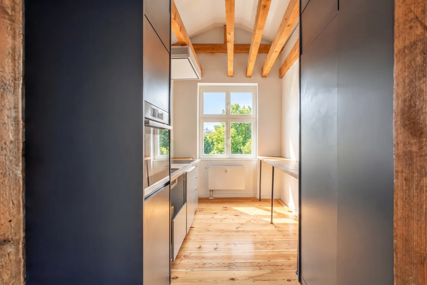 A narrow kitchen with light wood floors, white walls, and exposed wood beams. A window overlooks green trees. Dark cabinets frame the view.
