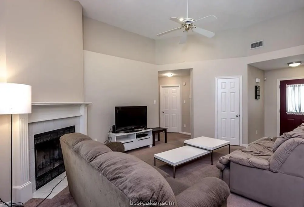 Living room with brown sofas, white coffee tables, TV, and fireplace. A ceiling fan hangs above. Neutral walls and carpet.