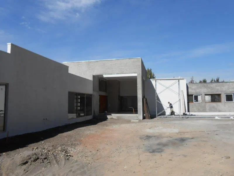 Exterior of a modern, gray stucco house under a blue sky. A man works on scaffolding near the white wall. Windows and a red door are visible.