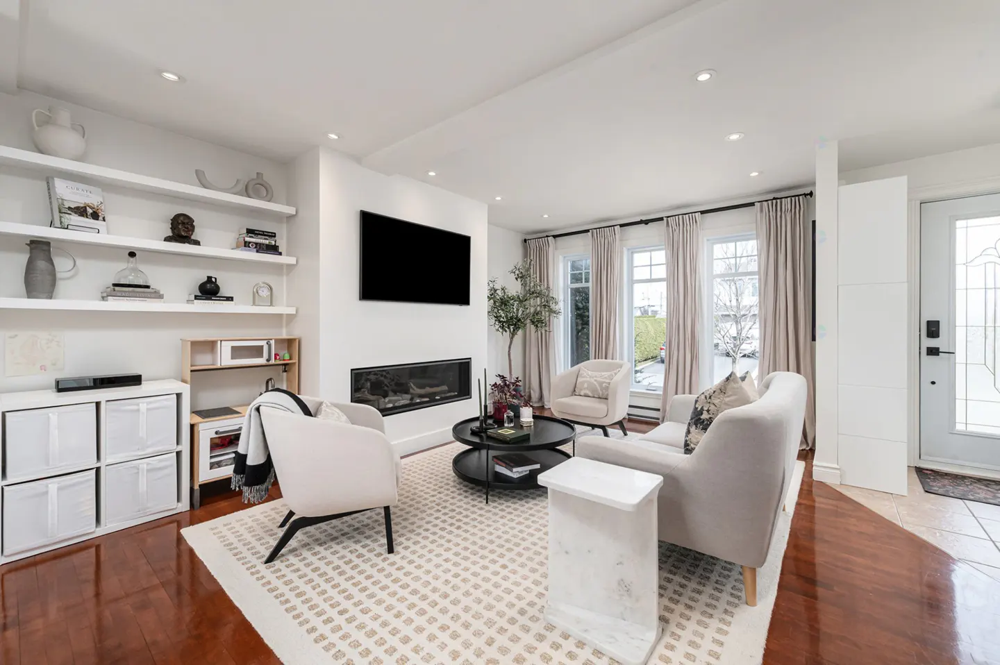 Bright living room with white walls, wood floors, and neutral furniture. A black TV hangs above a modern fireplace. Shelves display decor.