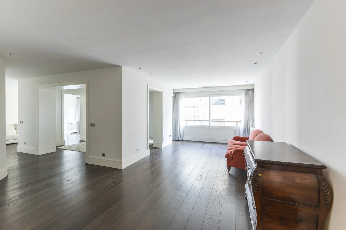 Bright, modern apartment interior with dark wood floors, white walls, and a large window. A red sofa and wooden dresser add color.