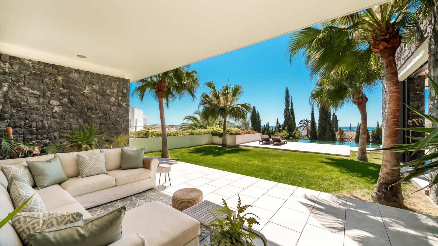 Outdoor patio with a cream sectional sofa, stone wall, and view of a green lawn, palm trees, and blue sky.
