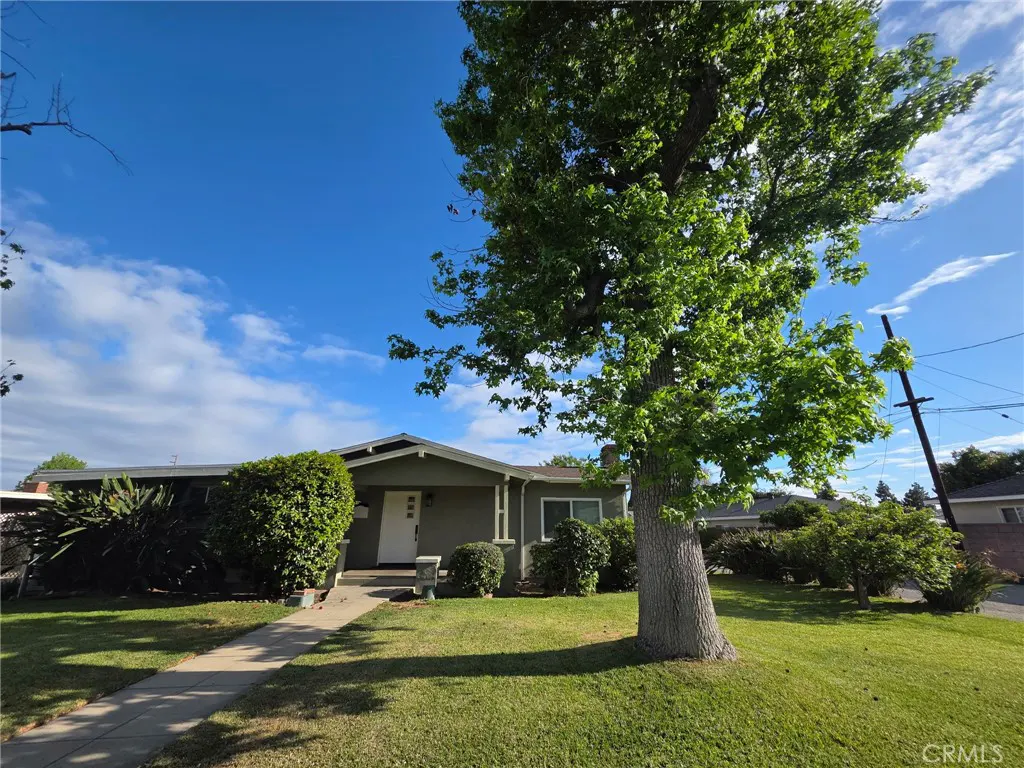 A single-story house with a green lawn, a walkway to the white front door, and a large tree in the yard. Blue sky with clouds.