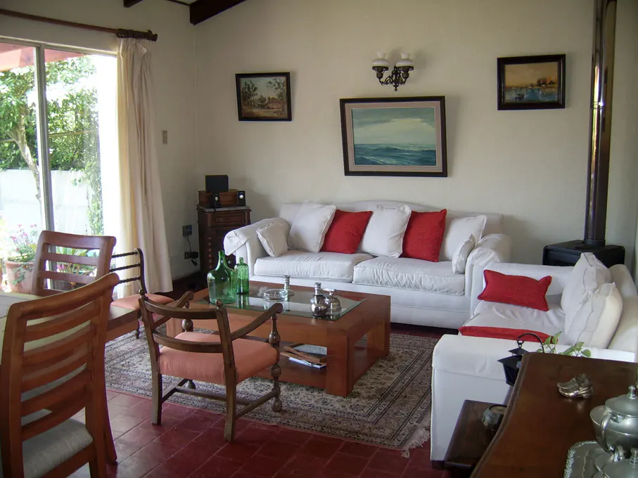 Cozy living room with white sofas, red pillows, wood furniture, and landscape paintings on the wall. A large window lets in natural light.