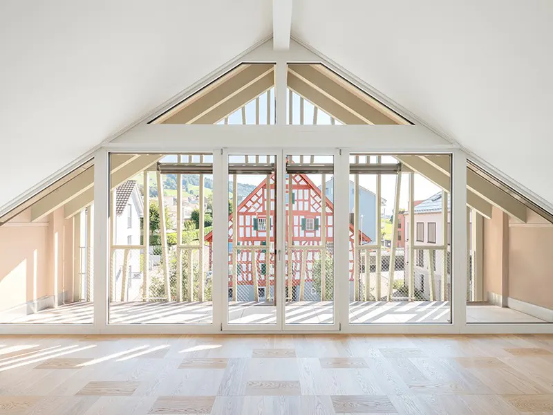 Attic room with white walls, light wood floors, and large windows. Outside, a red and white timber-framed house is visible.