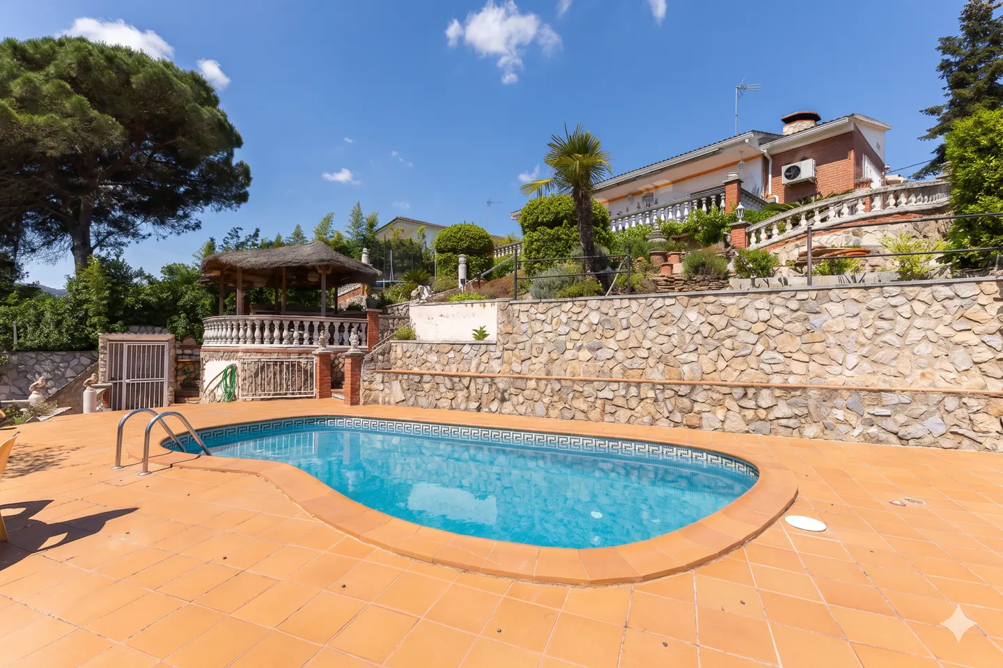 Outdoor pool with terracotta tiles, metal ladder, and gazebo. Stone wall leads to a house on a hill under a blue sky.