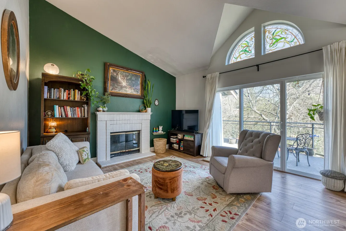 Living room with green accent wall, white fireplace, and large windows leading to a balcony. A beige sofa and armchair sit on a floral rug.