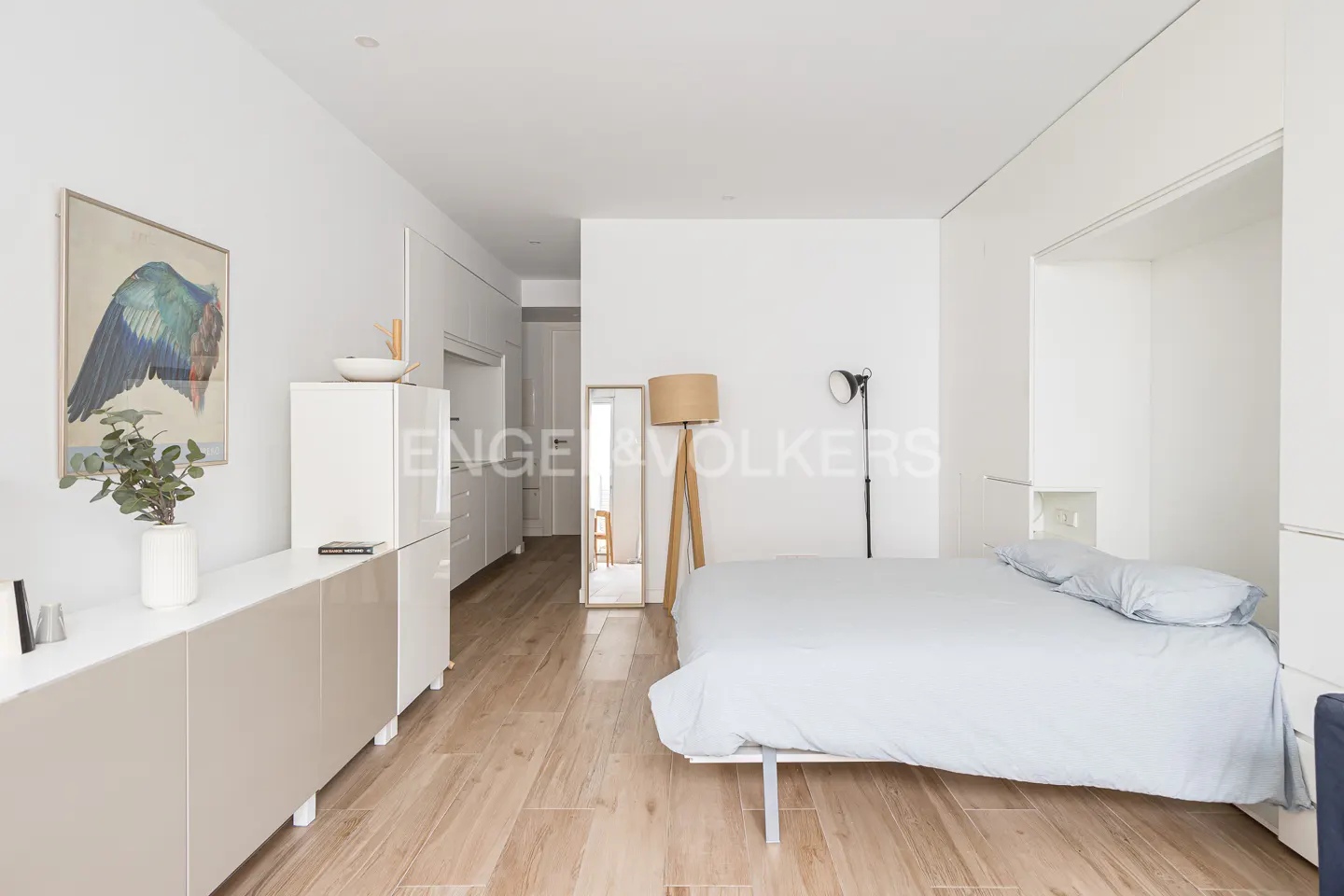 Bright studio apartment with wood floors, white walls, and a light blue bed. A tall mirror and modern lamps add to the minimalist design.